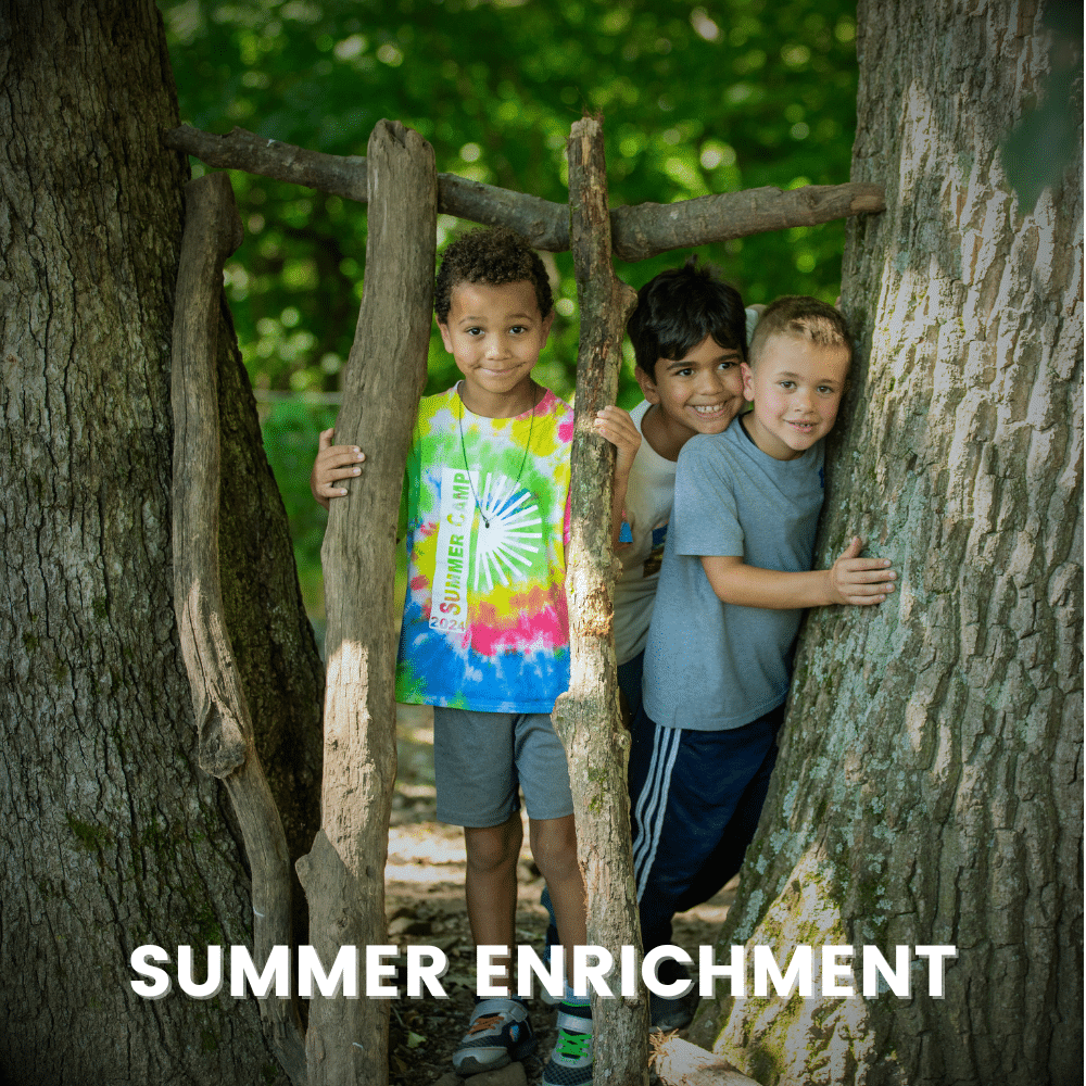 Three small children stand outside between two large trees